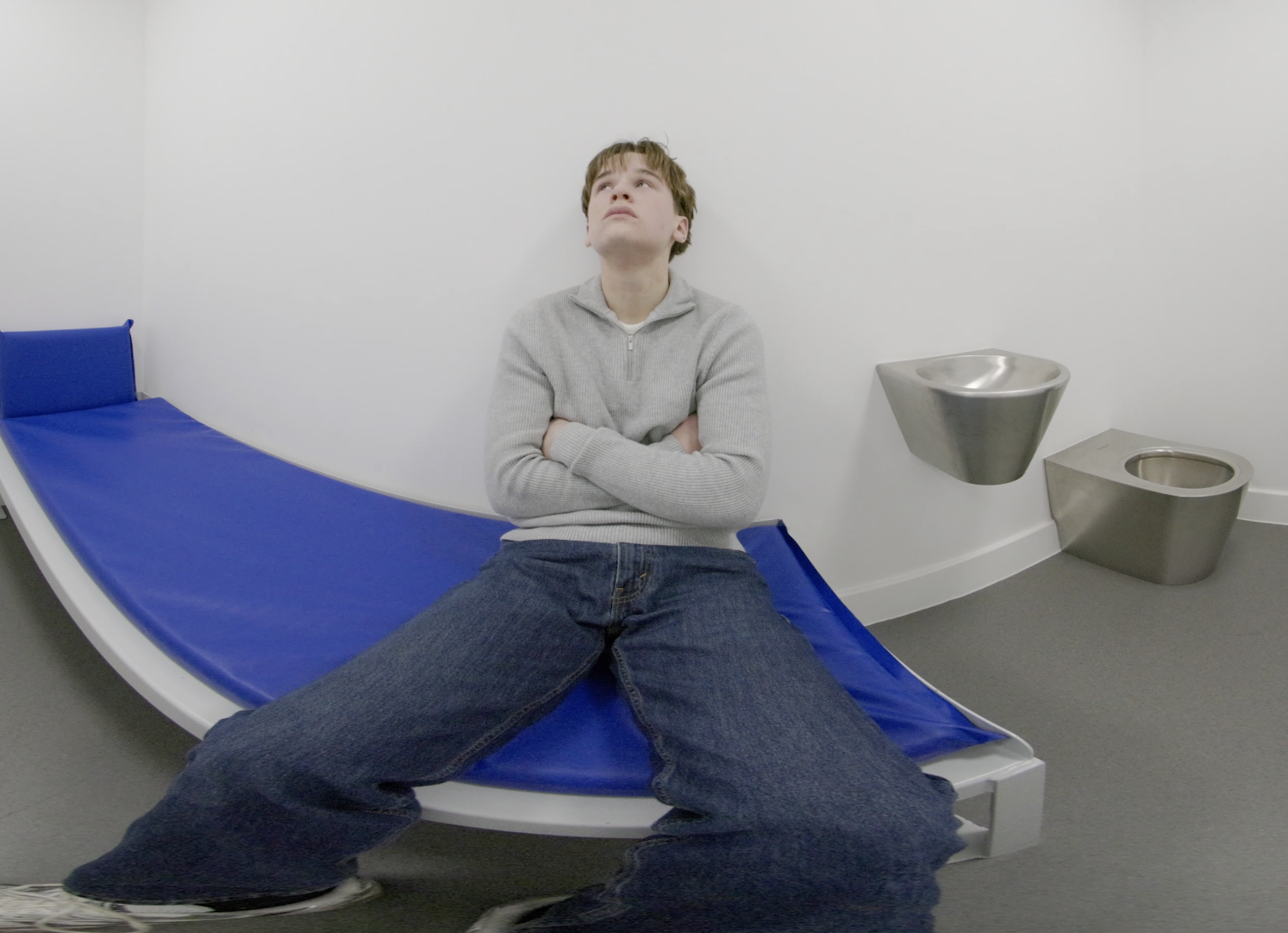 Still from the 3D 180° film: a teenage boy sitting with folded arms on a bed in a custody cell